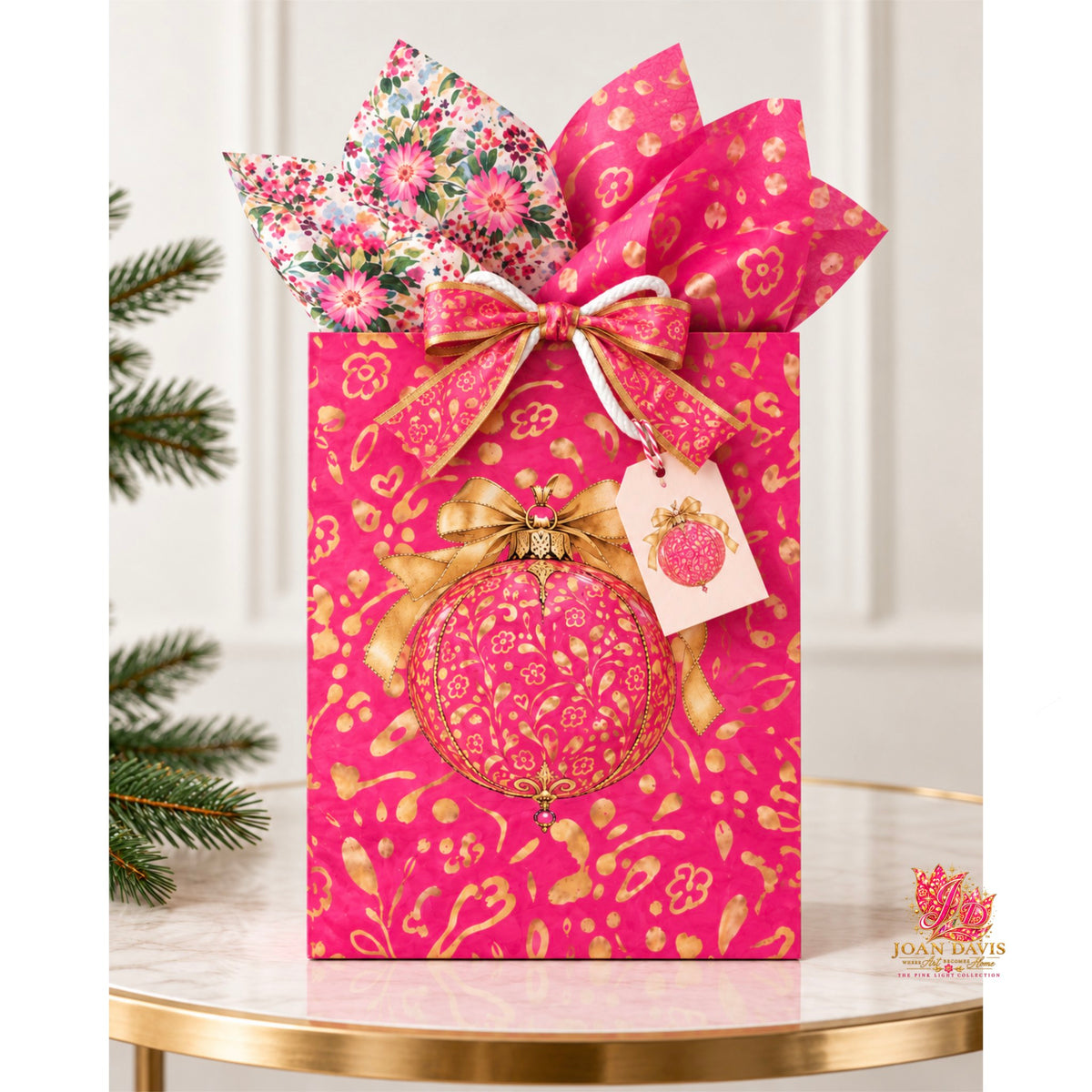 A pink gift bag with gold ornamental patterns, tied with a large pink bow and a smaller floral ribbon bow. A decorative pink ornament and a small tag are attached, and the bag is styled on a marble table with a gold edge, with pine branches at the side.